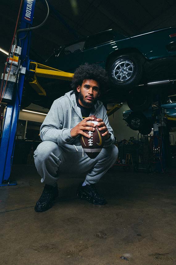 Dante Moore wearing Speedway hoodie and pants. He's holding a football with several cars in the background.