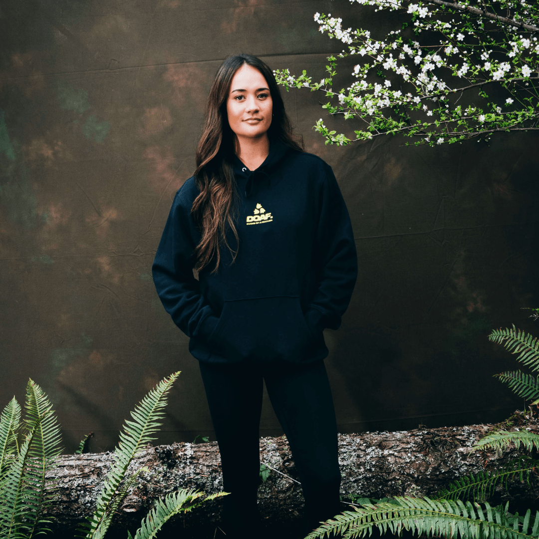 Kiara Romero wearing a black DOAF hoodie with a logo, standing outdoors with greenery around