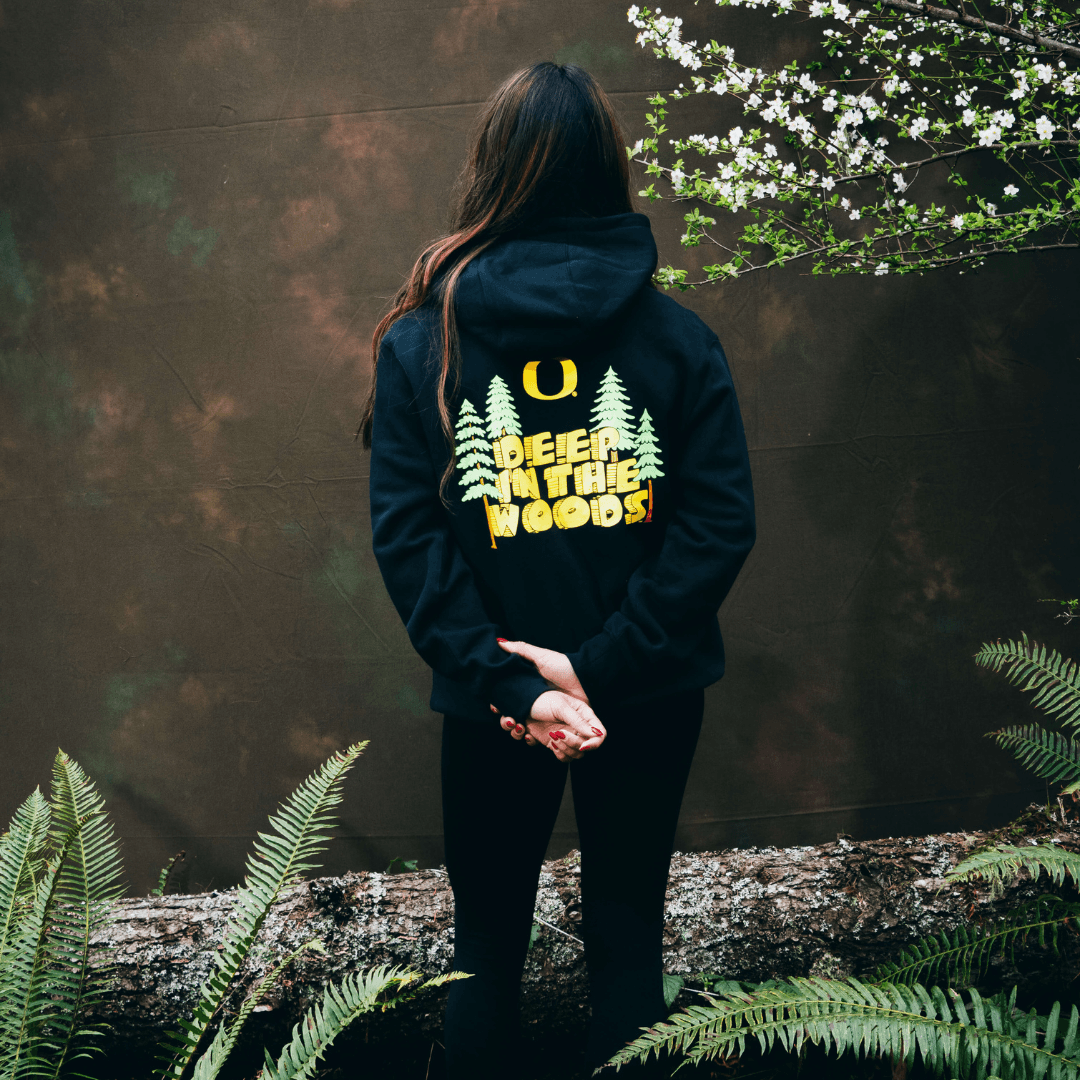 Kiara Romero wearing a black hoodie with 'Deer in the Woods' text and tree design, standing in a forest setting.