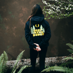 Kiara Romero wearing a black hoodie with 'Deer in the Woods' text and tree design, standing in a forest setting.