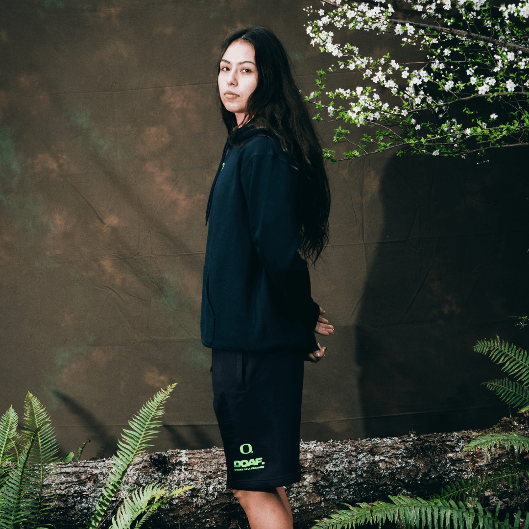 Ari Long wearing a dark outfit with 'DOAF' branding, standing against a textured wall with greenery.