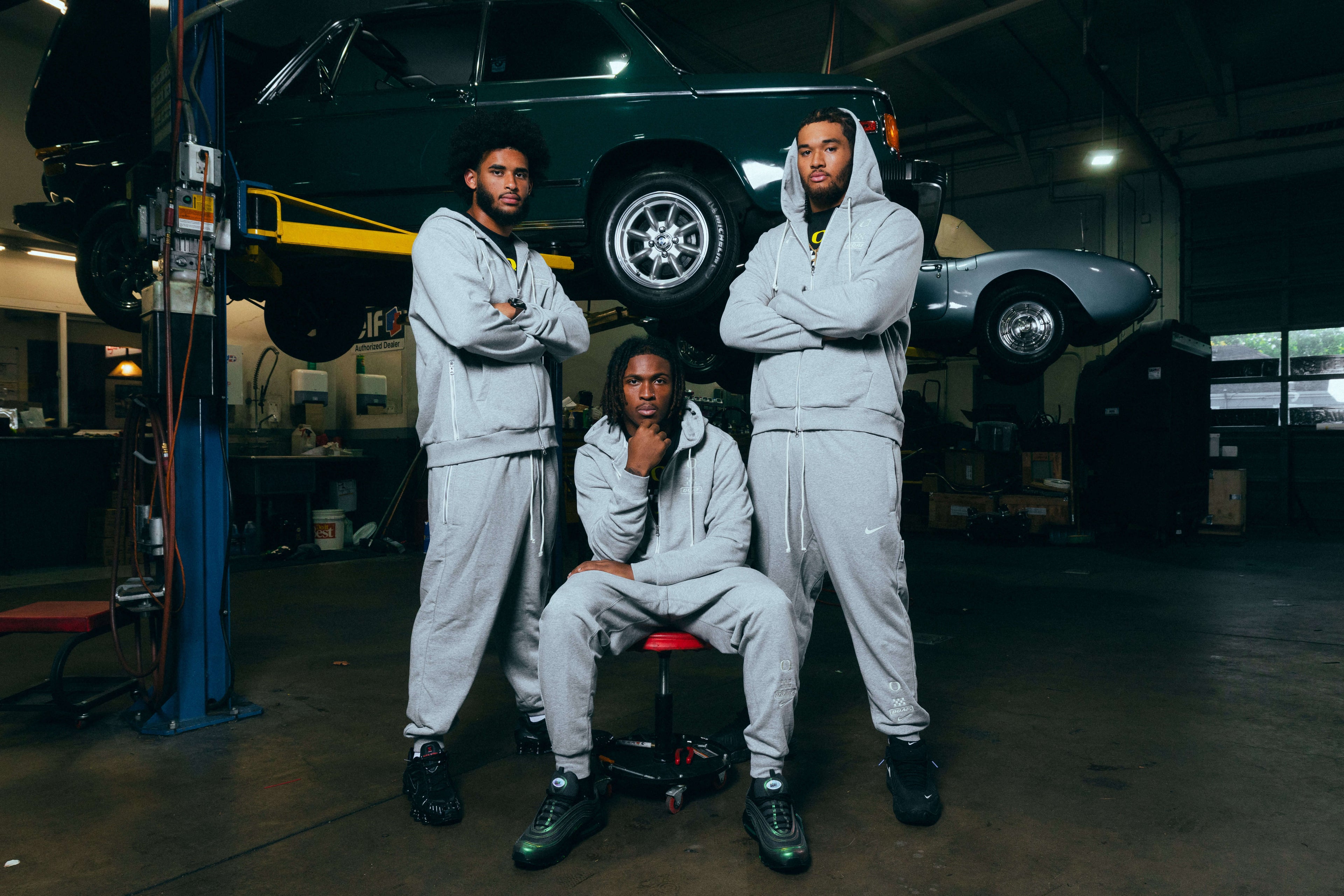 Dante Moore, Dakorien Moore, and Matayo Uiagalelei in matching outfits standing in a garage with cars in the background. They are wearing grey DOAF Speedway sweatshirts and pants.