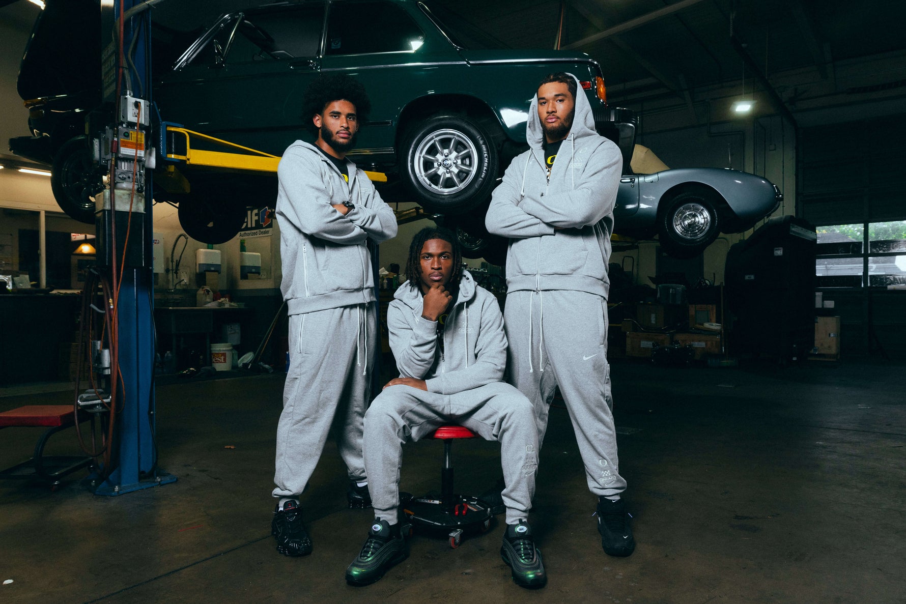 Dante Moore, Dakorien Moore, and Matayo Uiagalelei in DOAF Speedway Hoodies, Pants and Tees standing in a garage with cars in the background