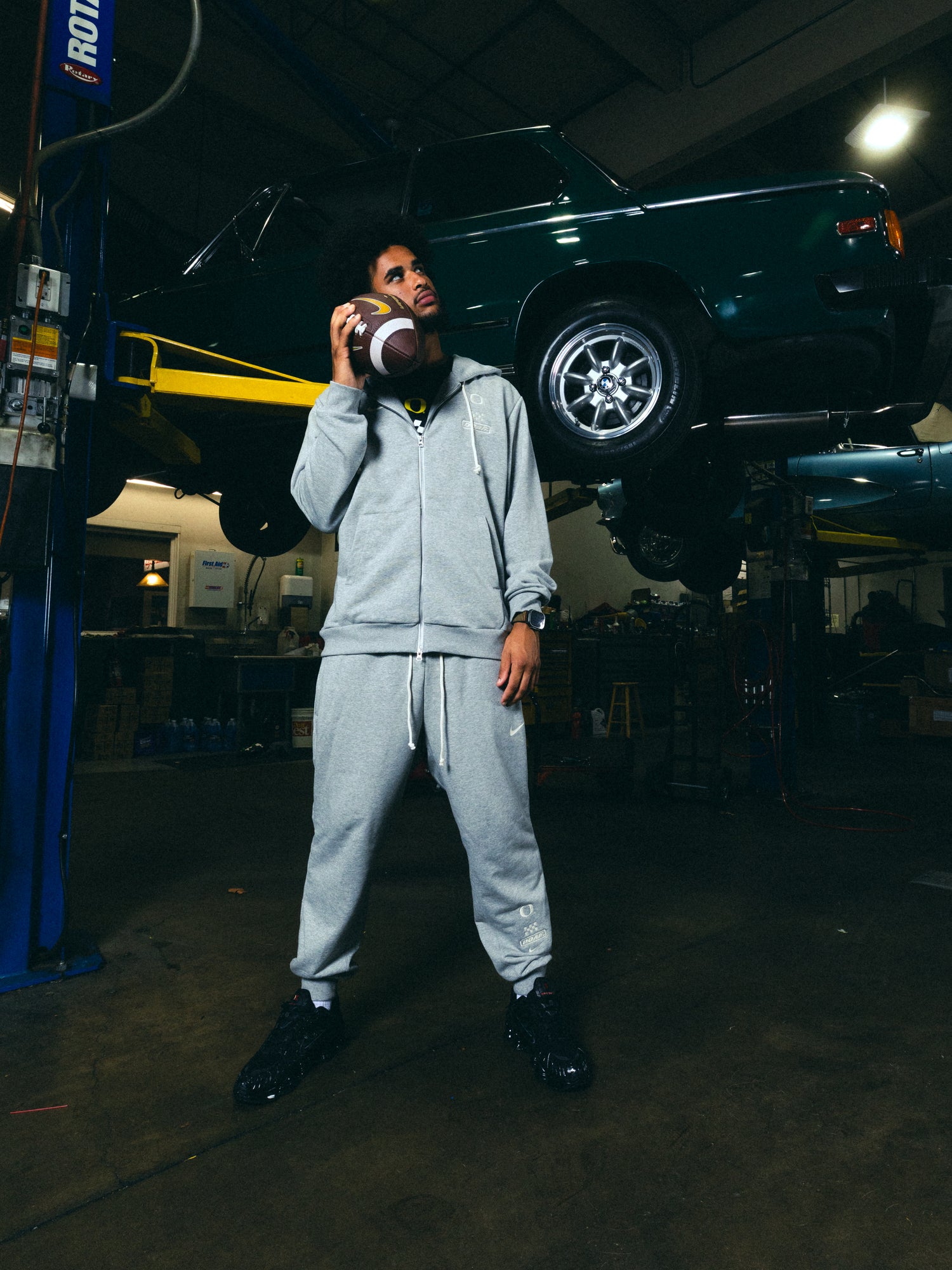 Dante Moore in grey Speedway hoodie and pants holding a football in a garage with a vehicle in the background