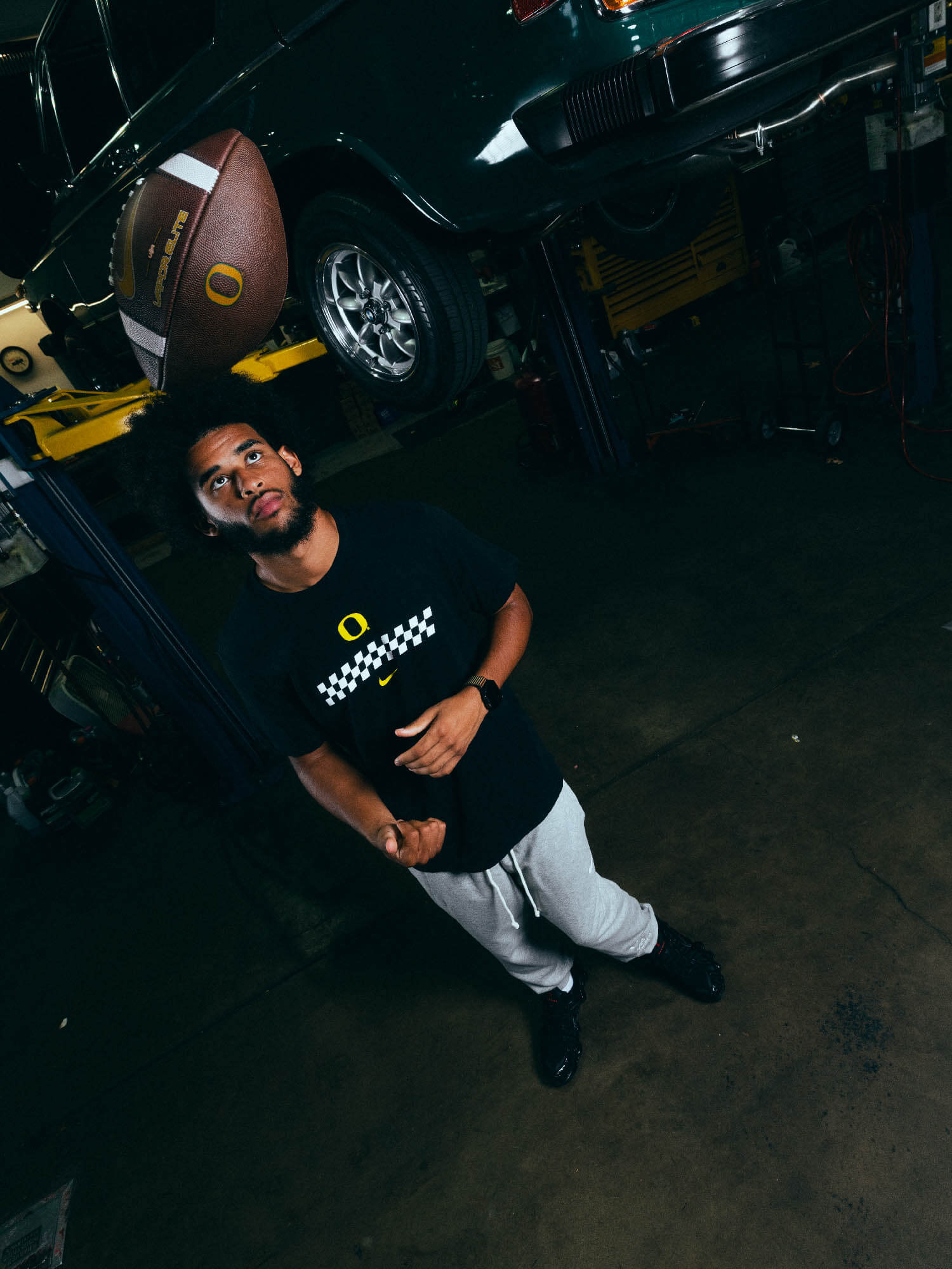 Dante Moore standing in a garage with a car and football in the background. He is wearing DOAF Speedway Tee and Pants.