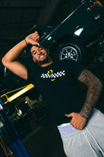 Matayo Uiagalelei, wearing a black Speedway Tee and grey Speedway pants, is posing in a garage with a car in the background.
