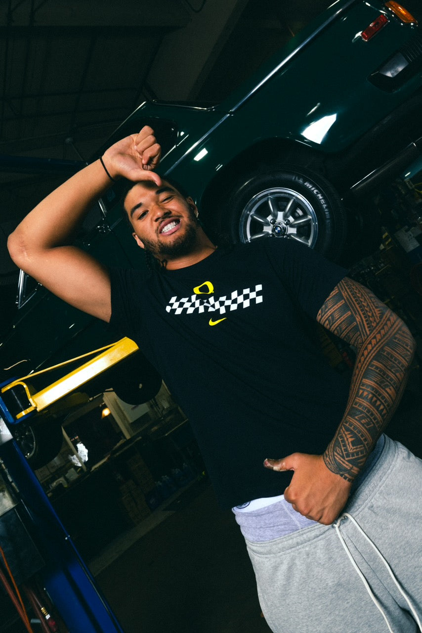 Matayo Uiagalelei, wearing a black Speedway Tee and grey Speedway pants, is posing in a garage with a car in the background.