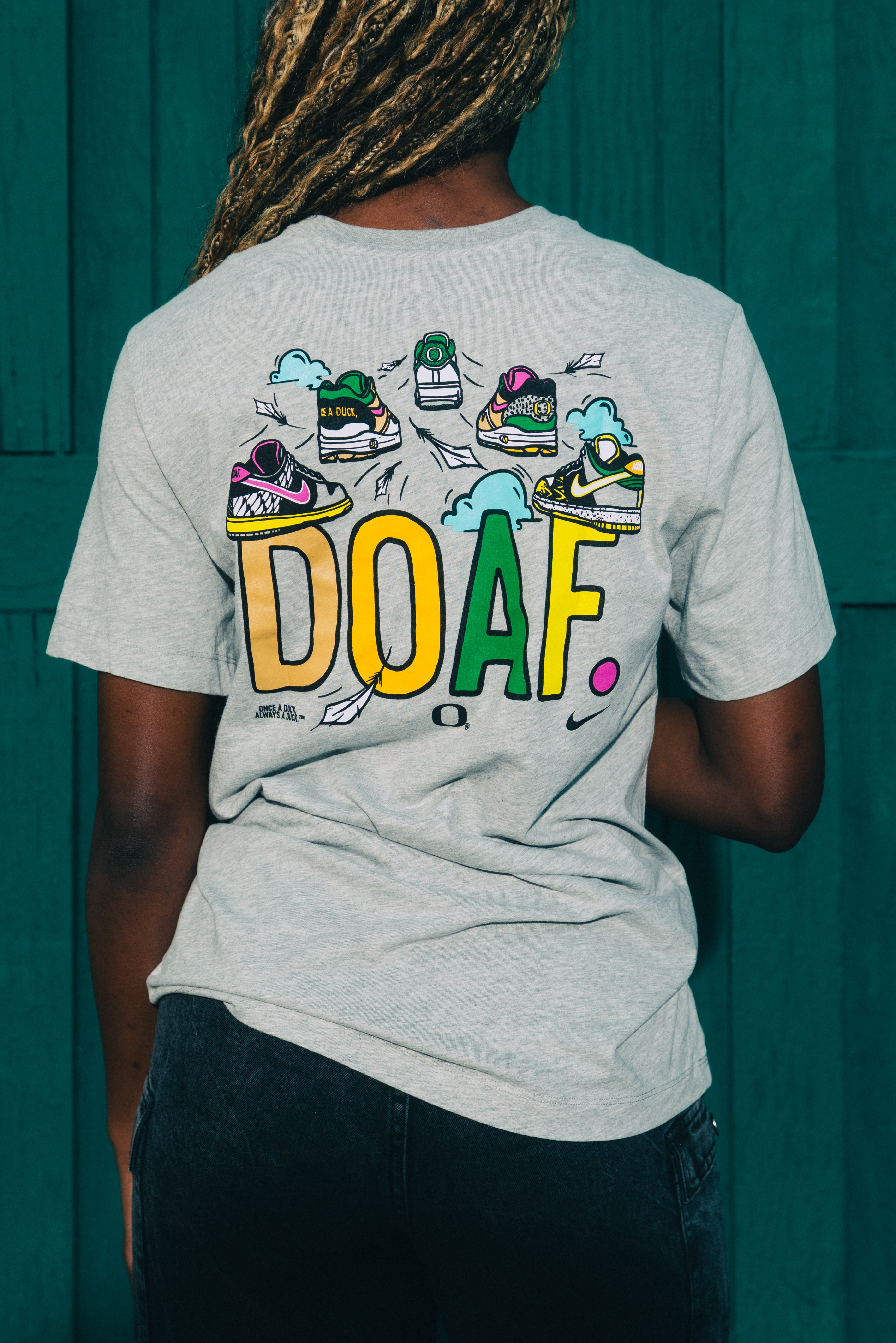 Amina Muhammad wearing a gray t-shirt with colorful sneaker graphics and 'DOAF' text against a teal background