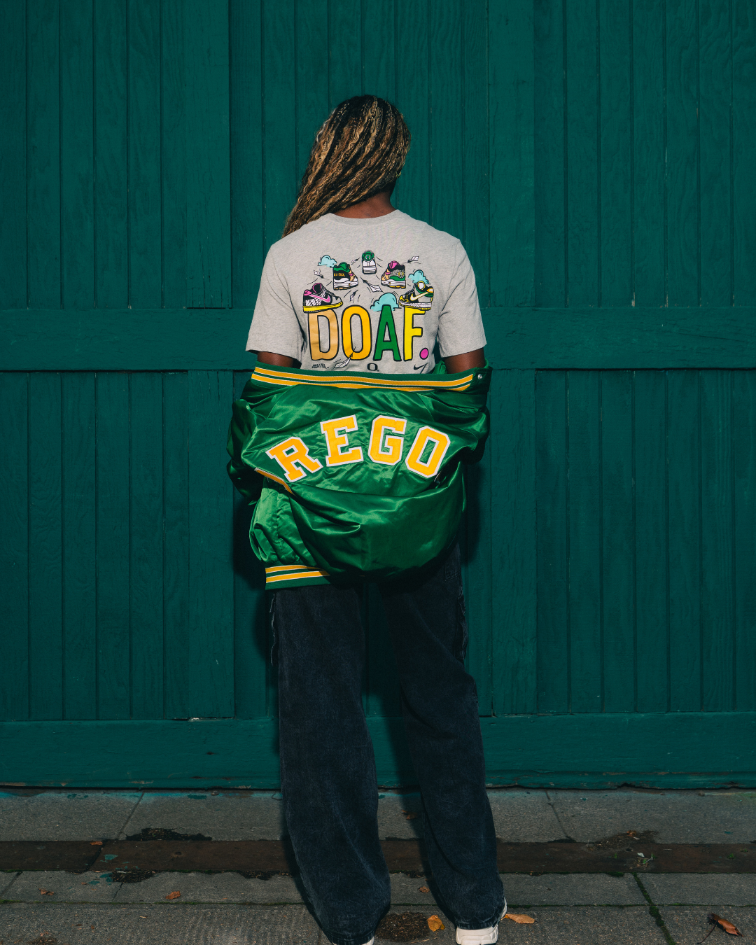 Amina Muhammad wearing a DOAF Sneaker Tee with a green Oregon jacket, standing against a teal wall.