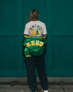Amina Muhammad wearing a DOAF Sneaker Tee with a green Oregon jacket, standing against a teal wall.