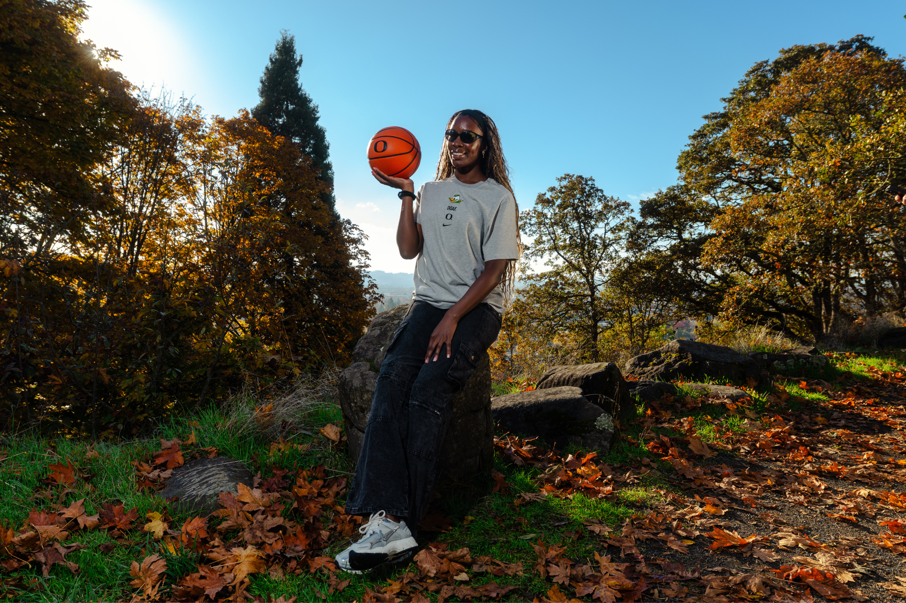 Amina Muhammad holding a basketball outdoors in a forest setting with trees and fallen leaves. She is wearing a grey DOAF tee
