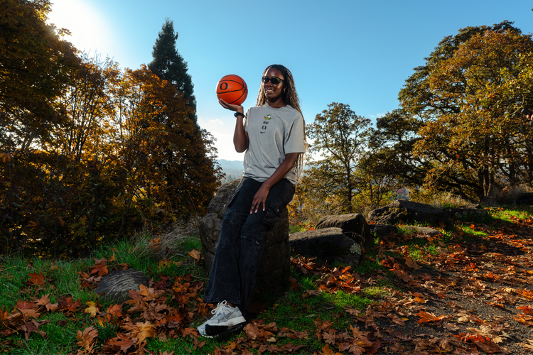Amina Muhammad holding a basketball outdoors in a forest setting with trees and fallen leaves. She is wearing a grey DOAF tee