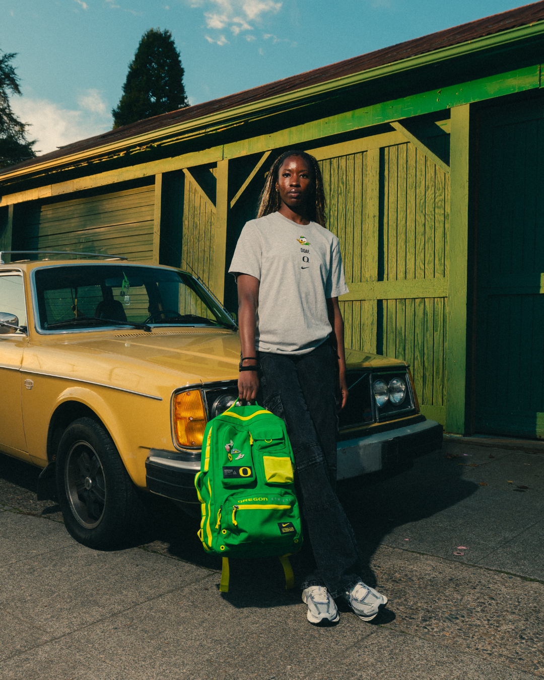 Amina Muhammad standing next to a yellow car with a green backpack, wearing a grey DOAF sneaker t-shirt and dark pants.