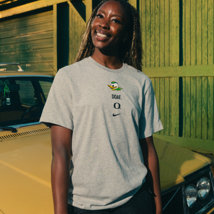 Amina Muhammad wearing a gray DOAF branded t-shirt with logos standing next to a yellow car.