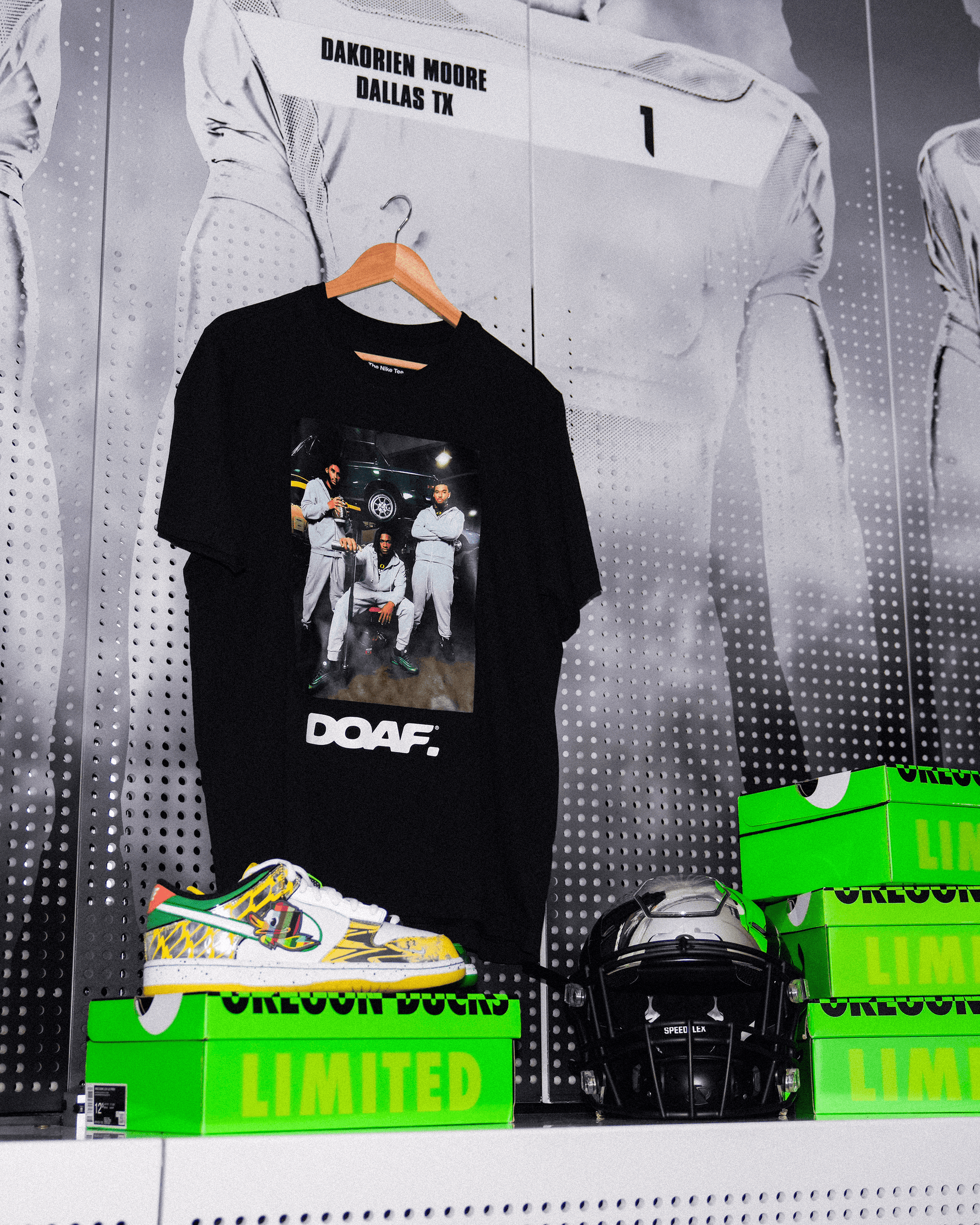 DOAF Tee featuring Dante Moore, Dakorien Moore, and Matayo Uiagalelei hanging up on Dakorien's locker. There is a football helmet and sneakers stacked on DOAF shoe boxes.