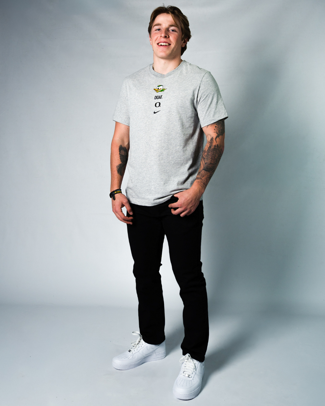 Atticus Sappington wearing a gray t-shirt with DOAF logos, black pants, and white sneakers on a plain background