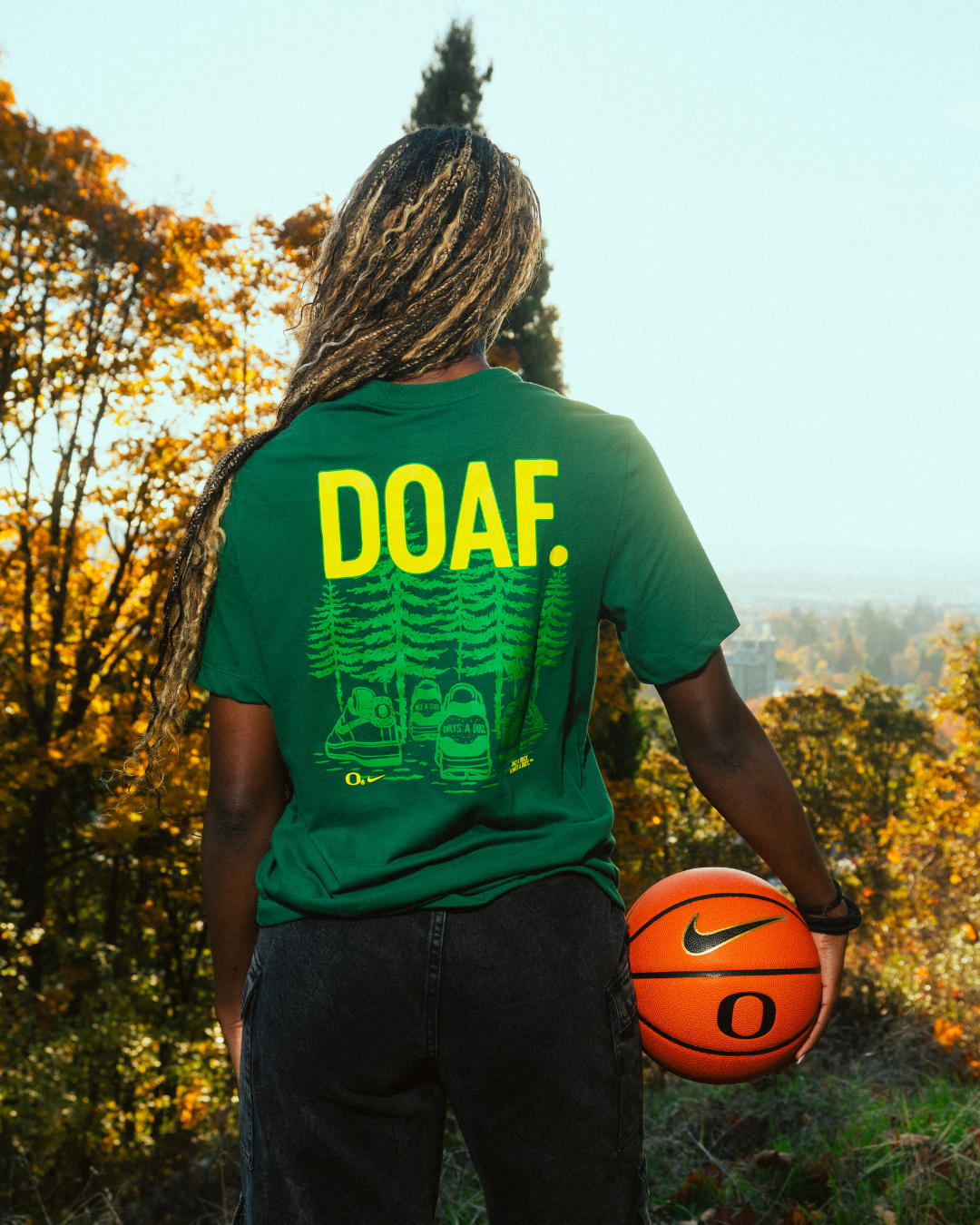 Amina Muhammad wearing a green t-shirt with sneaker illustrations and 'DOAF.' text, holding a basketball outdoors.