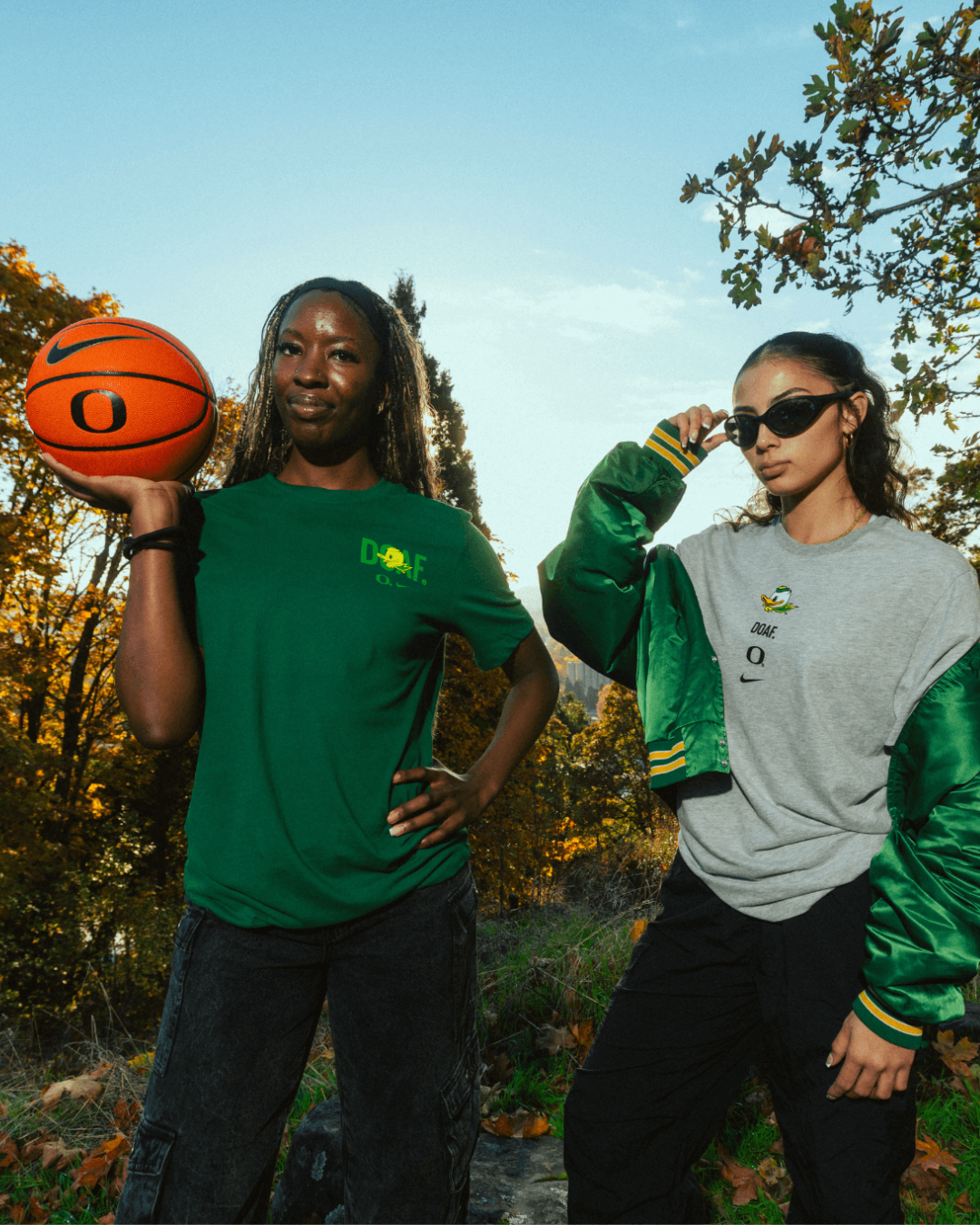 Amina Muhammad holding a basketball, wearing green DOAF tee. Katie Fiso wearing sunglasses and a green jacket with a grey DOAF tee.