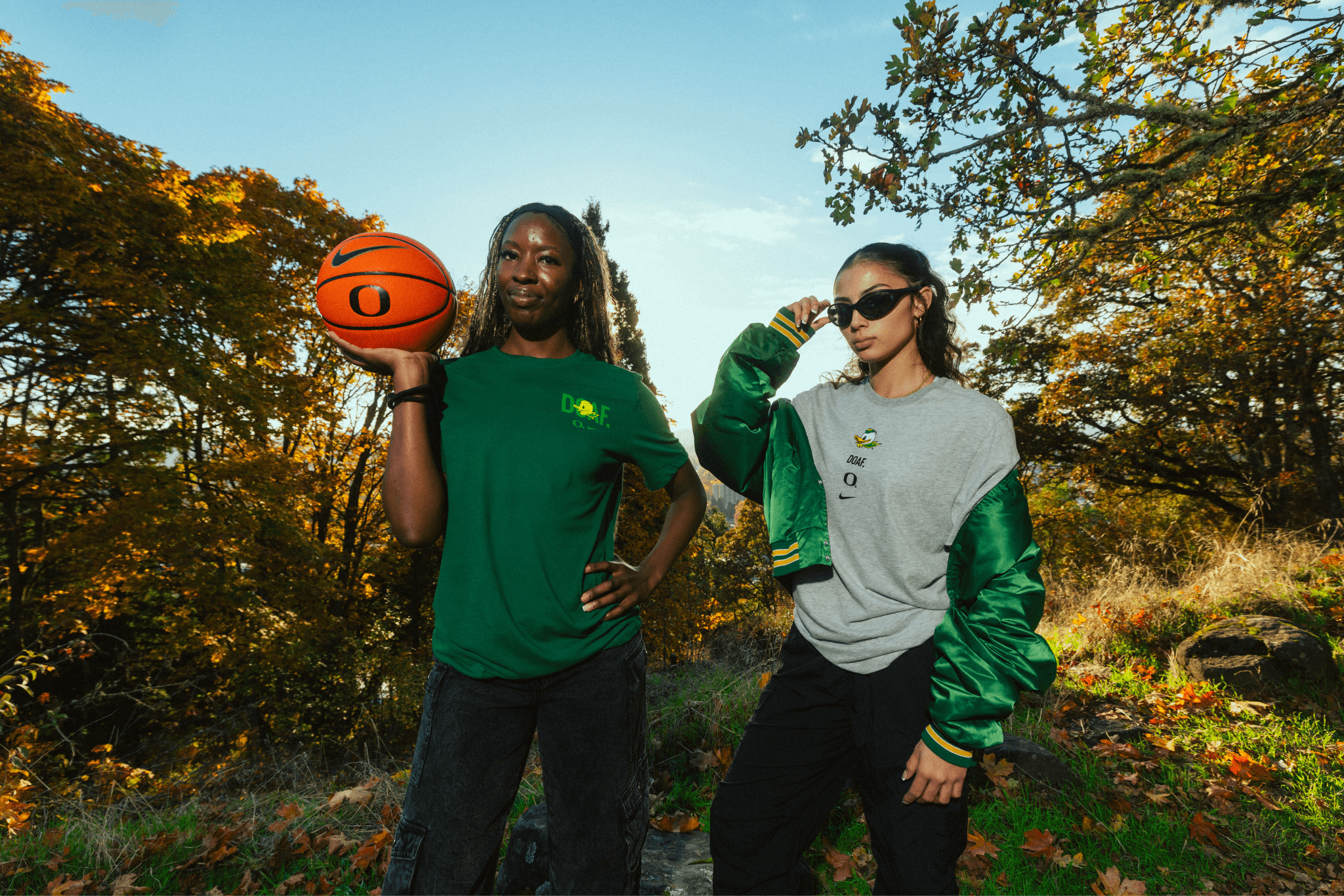 Amina Muhammad and Katie Fiso wearing DOAF sneaker tees standing outdoors with trees and a clear sky in the background