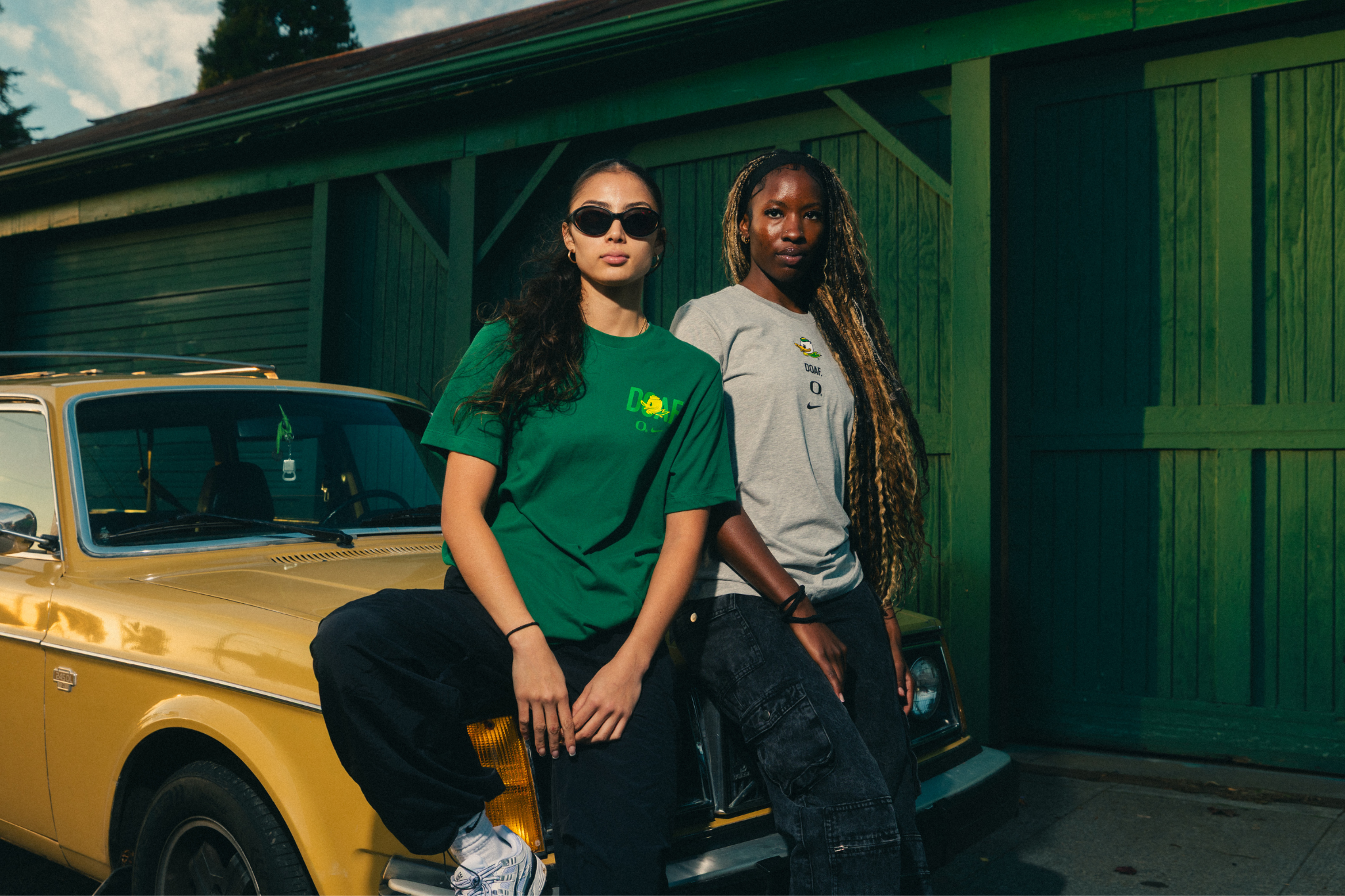 Amina Muhammad and Katie Fiso wearing DOAF Sneaker Tees and standing next to a yellow car in front of a green building.