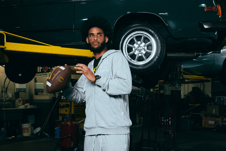 Dante Moore holding a football in a garage with a car in the background. He wearing DOAF Speedway Hoodie and Pants.