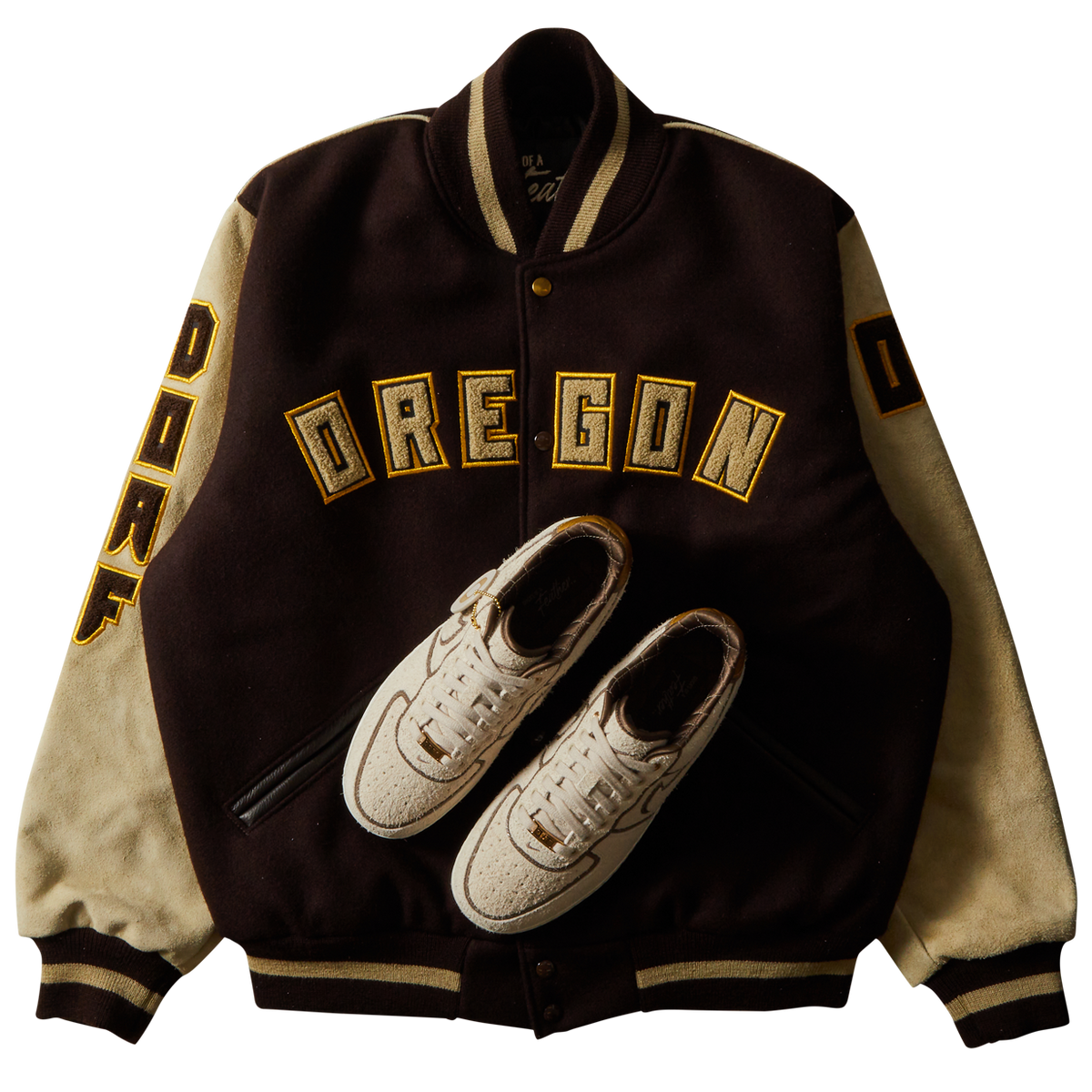 DOAF Varsity jacket with 'OREGON' text and shoe design on a white background. The sneakers reimagine the AF1 ’07 in a sophisticated combination of color and texture that manages to be both luxe and low-key at the same time. An ultra-premium high-texture suede upper in a creamy natural color.
The DOAF Varsity Jacket features a strong “OREGON” read across the chest and "DOAF" on the right sleeve. On the upper back, a metallic gold patch featuring the iconic “O” is stitched onto the jacket.