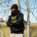 Person wearing a black hoodie with yellow text standing in front of a large metal structure.