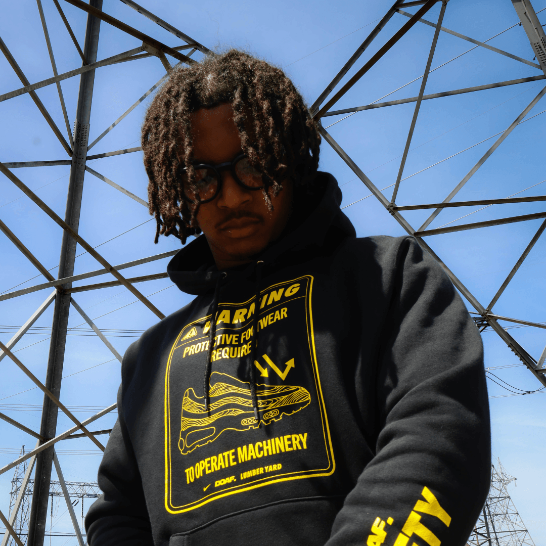 Person wearing a black hoodie with yellow text and graphics against a blue sky with metal structures.