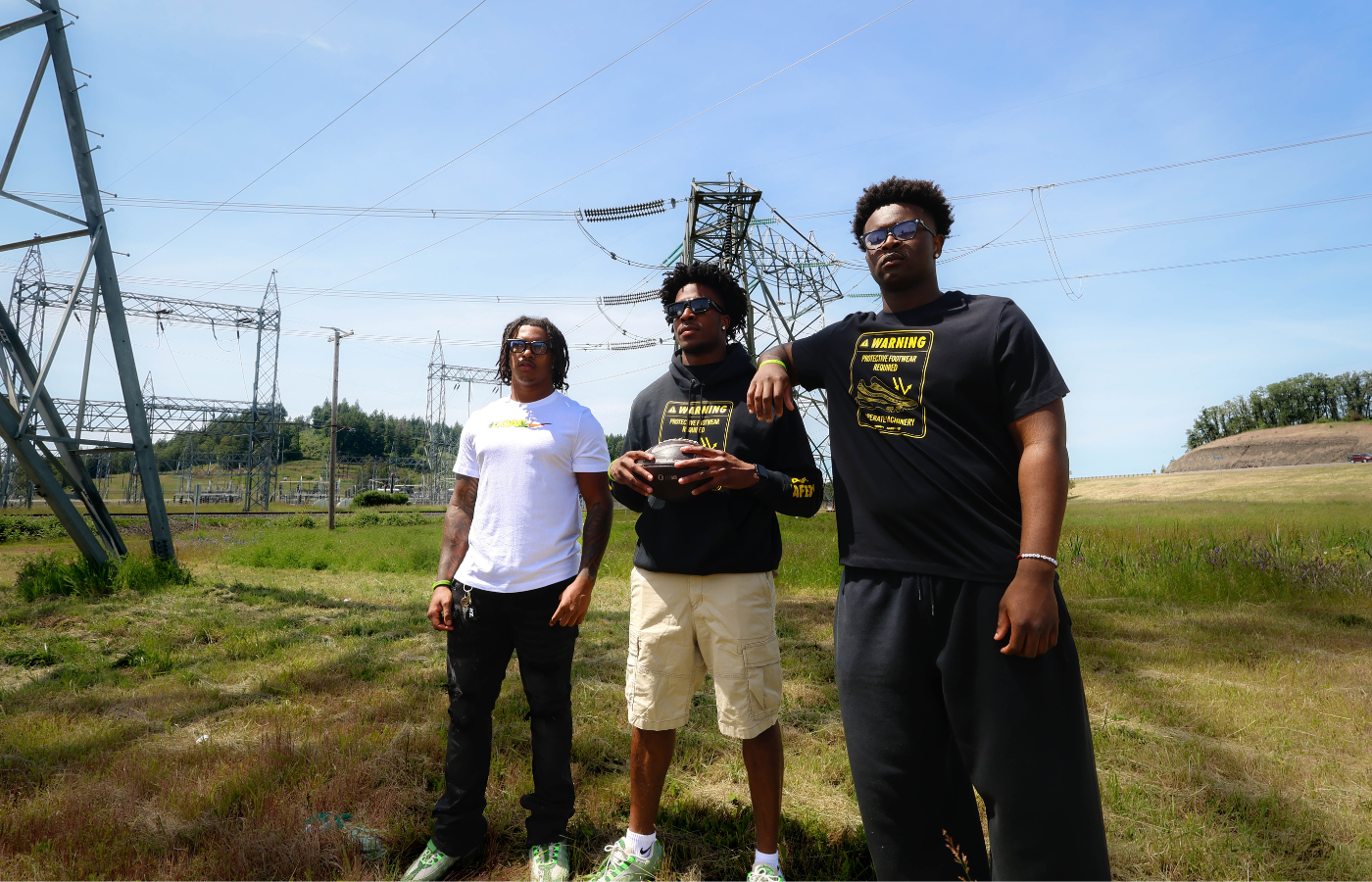 Three individuals standing in an open field with power lines and towers in the background.