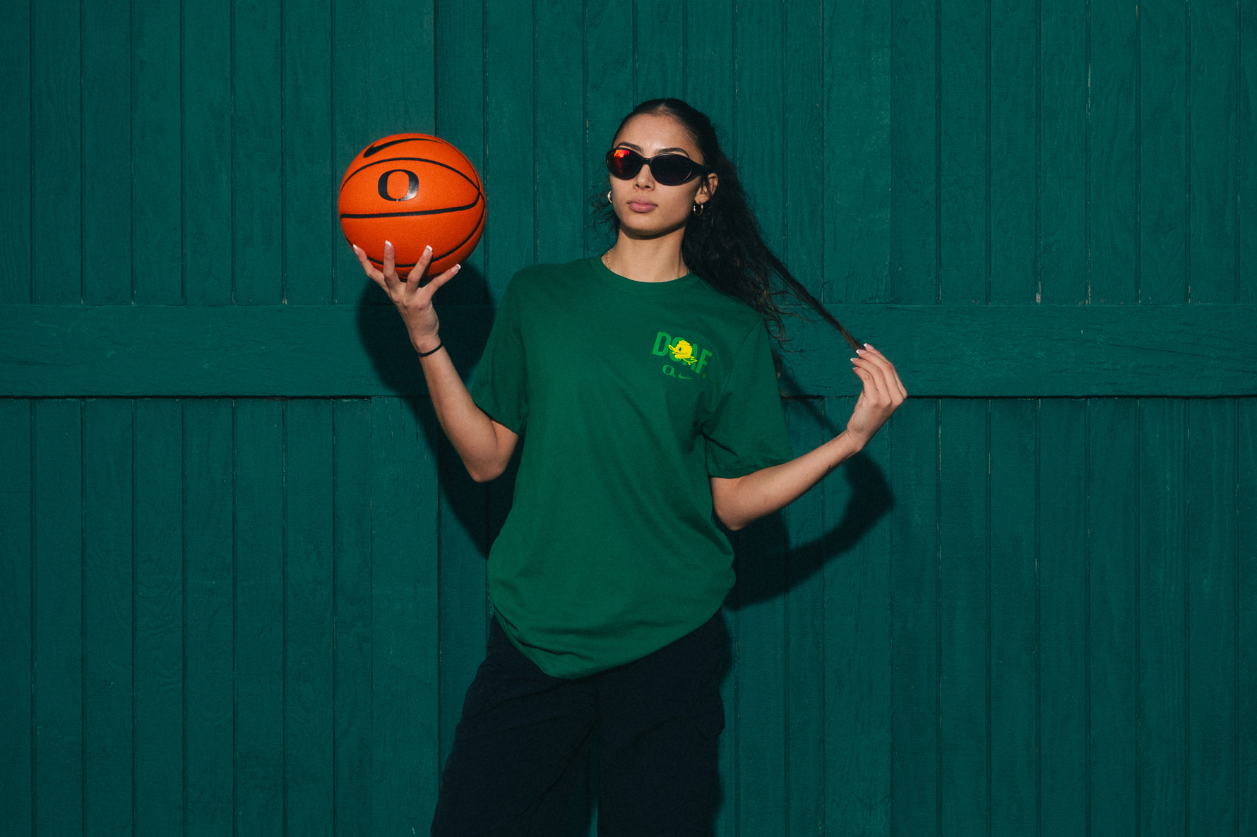 Katie Fiso holding a basketball against a green wall. She is wearing a green DOAF tee