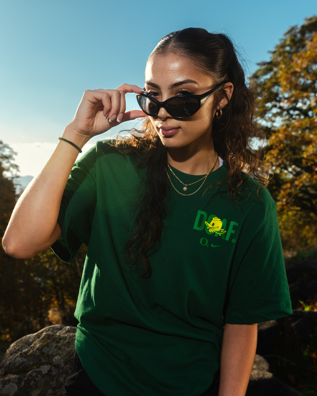 Katie Fiso wearing a green DOAF t-shirt with a logo, sitting outdoors with trees in the background