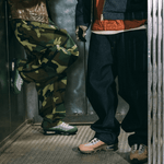 Two people wearing colorful pants and  DOAF sneakers in a dimly lit room.