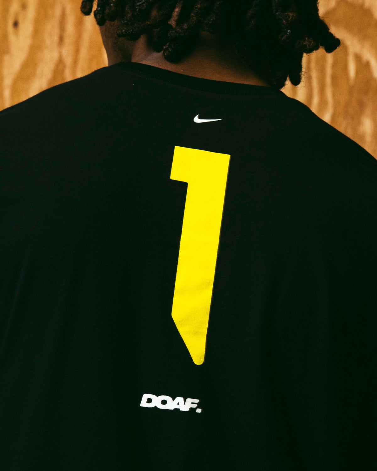 Black t-shirt with yellow number 1 and white logo on the back, worn by Dakorien Moore against a wooden wall.