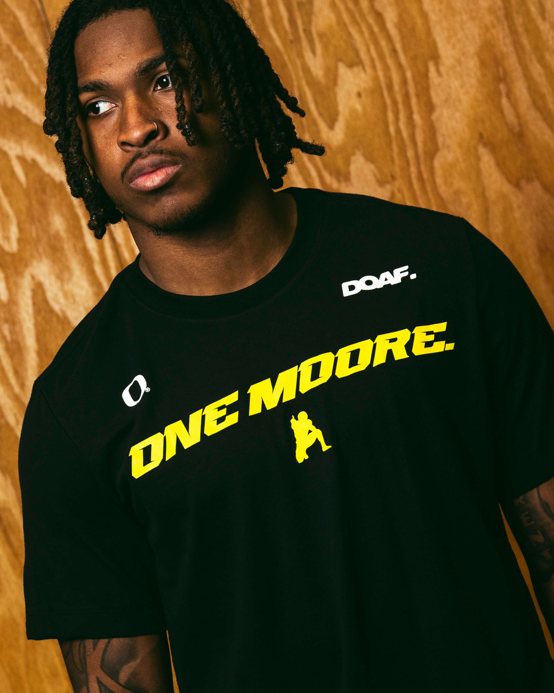 Dakorien Moore wearing a black t-shirt with 'ONE MOORE' text and logos on a wooden background
