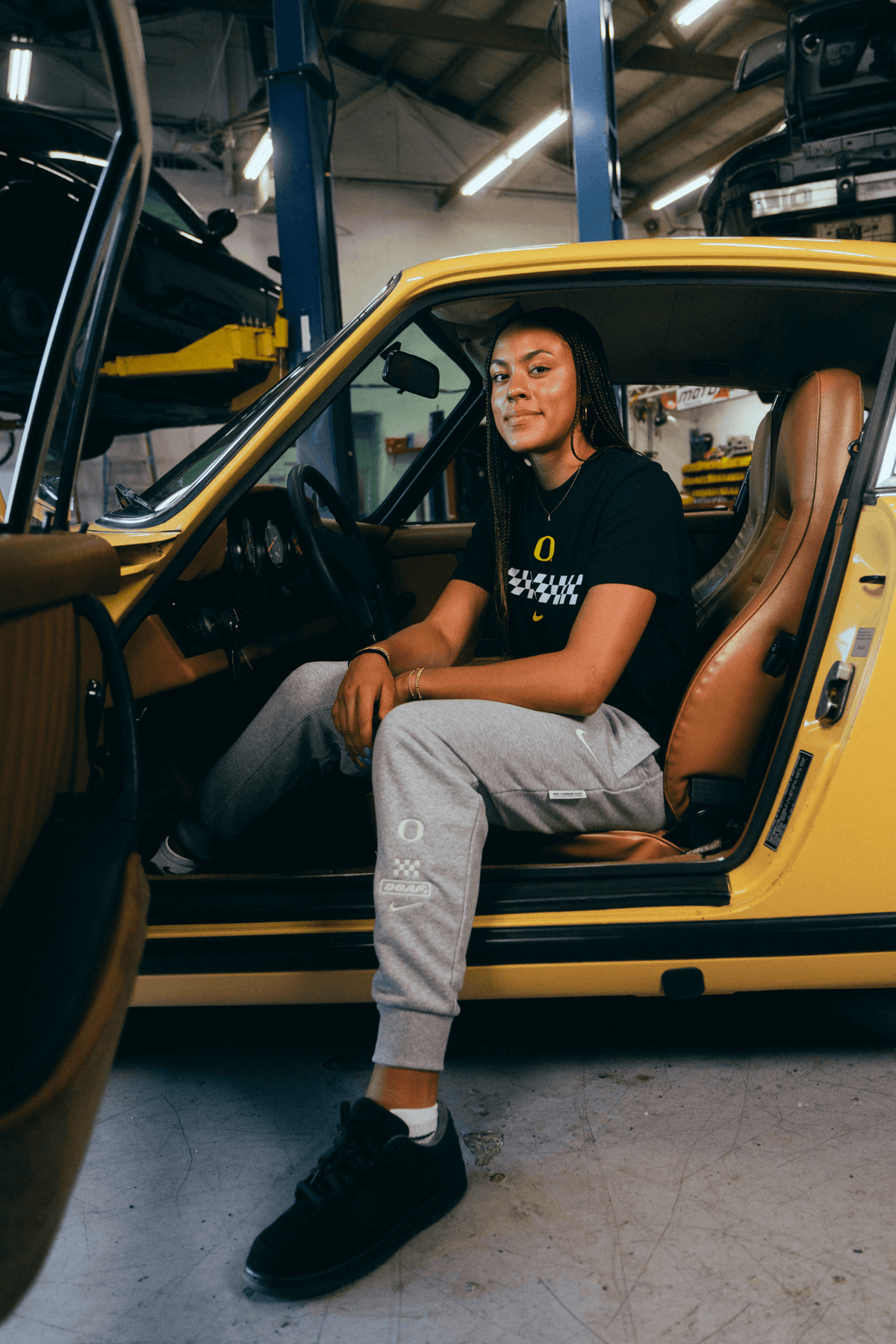 Sophia Bell sitting inside a yellow car in a garage. She's weearing a black DOAF Speedway t-shirt with a white checker graphic and grey Speedway sweat pants.