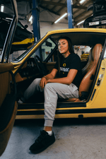 Sophia Bell sitting inside a yellow car in a garage. She's weearing a black DOAF Speedway t-shirt with a white checker graphic and grey Speedway sweat pants.