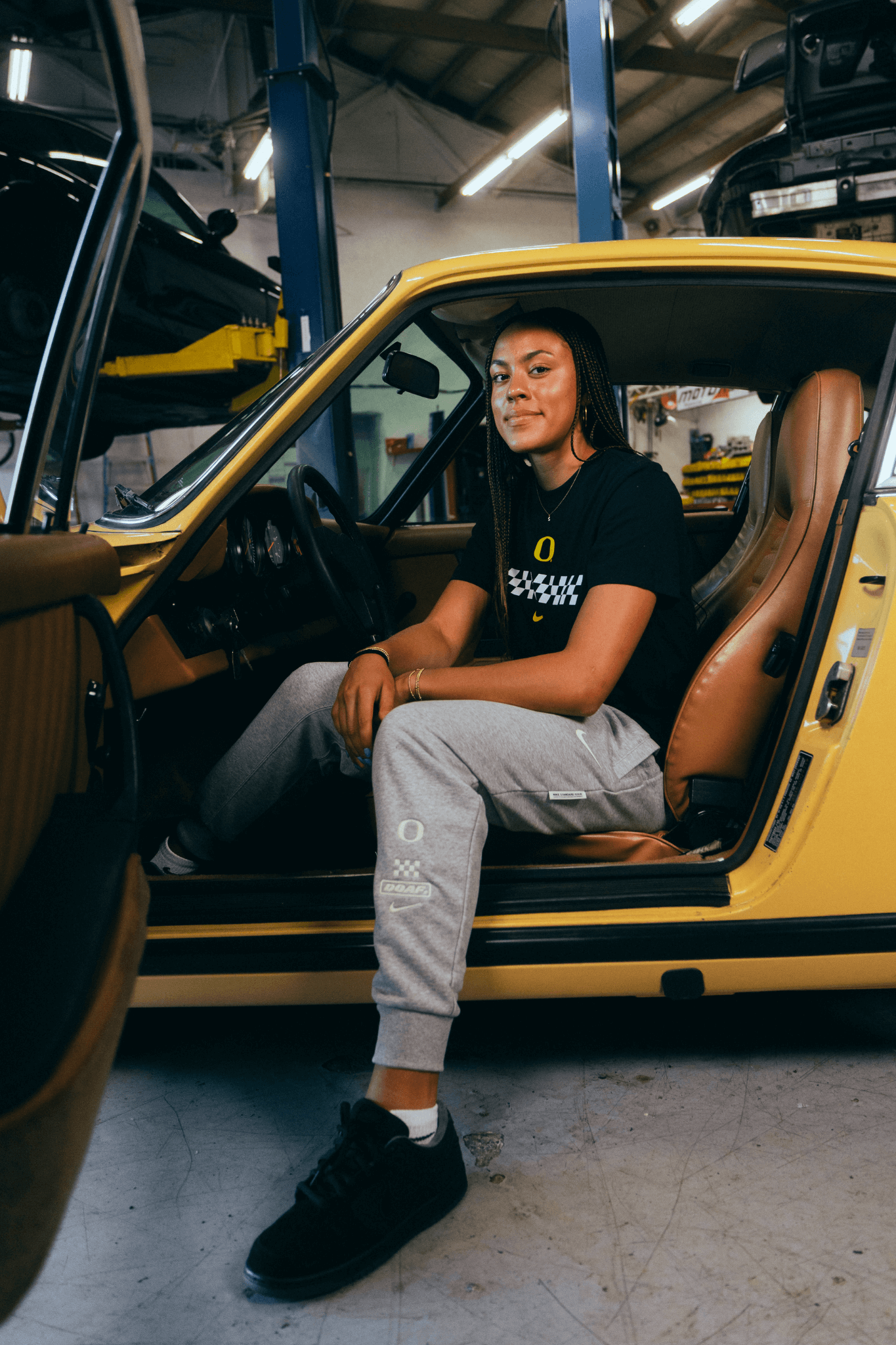 Sophia Bell sitting inside a yellow car in a garage. She's weearing a black DOAF Speedway t-shirt with a white checker graphic and grey Speedway sweat pants.