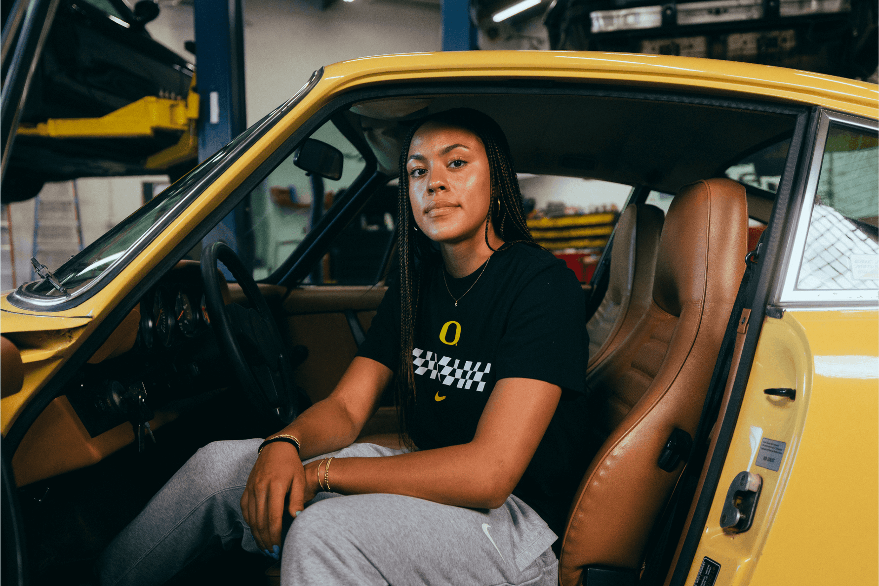 Sofia Bell sitting inside a yellow car in a garage. She is wearing a DOAF Speedway Tee and Pants.