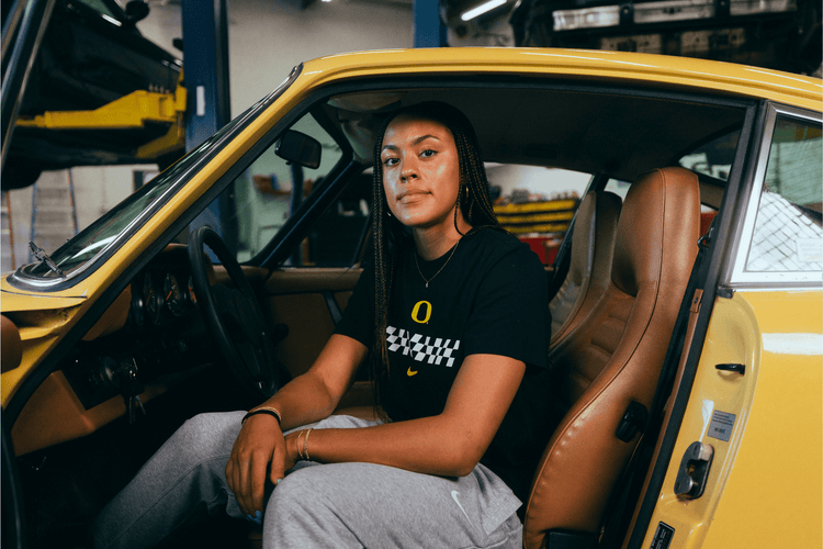 Sofia Bell sitting inside a yellow car in a garage. She is wearing a DOAF Speedway Tee and Pants.
