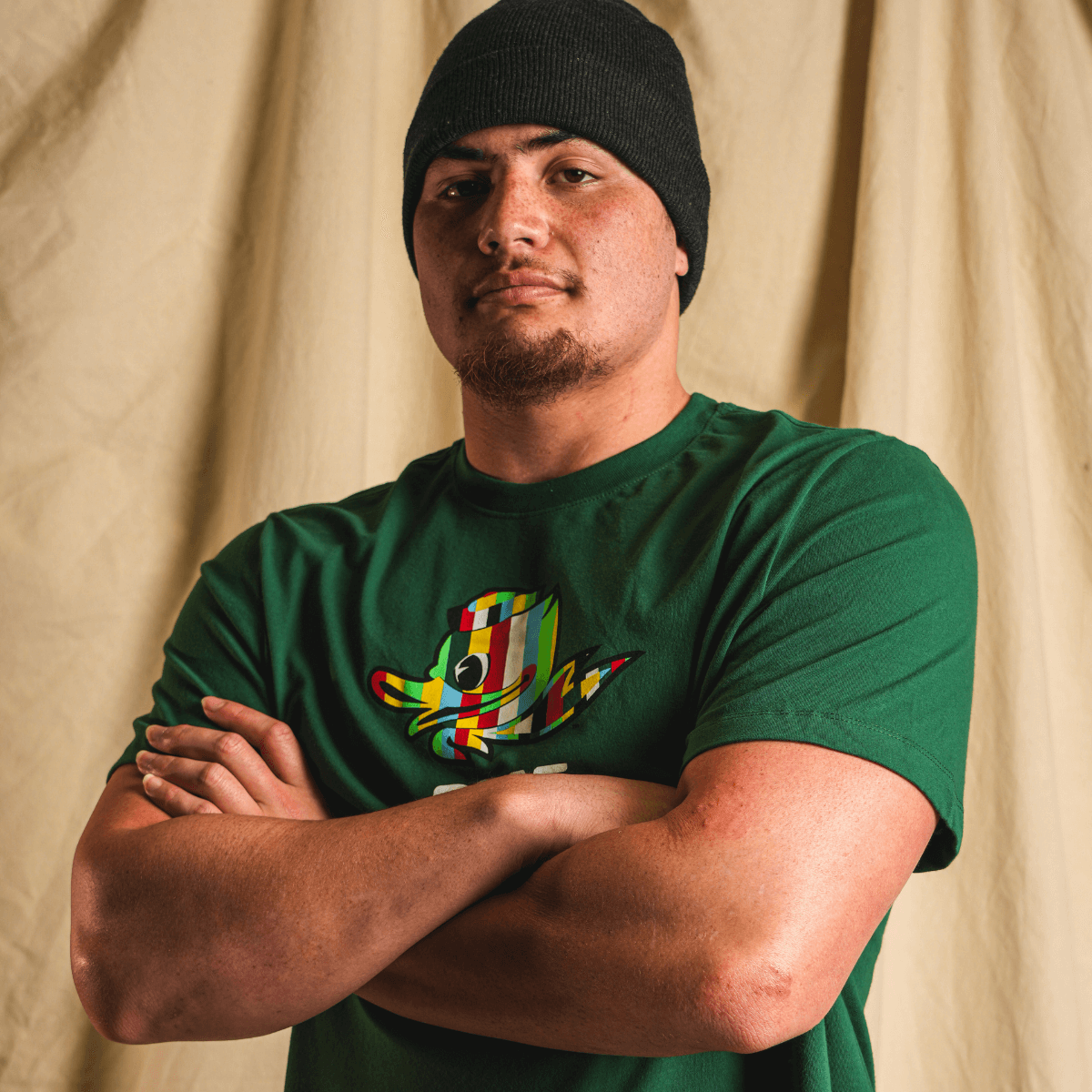 Teitum Tuioti wearing a green DOAF t-shirt with a colorful Duck graphic design against a beige curtain background