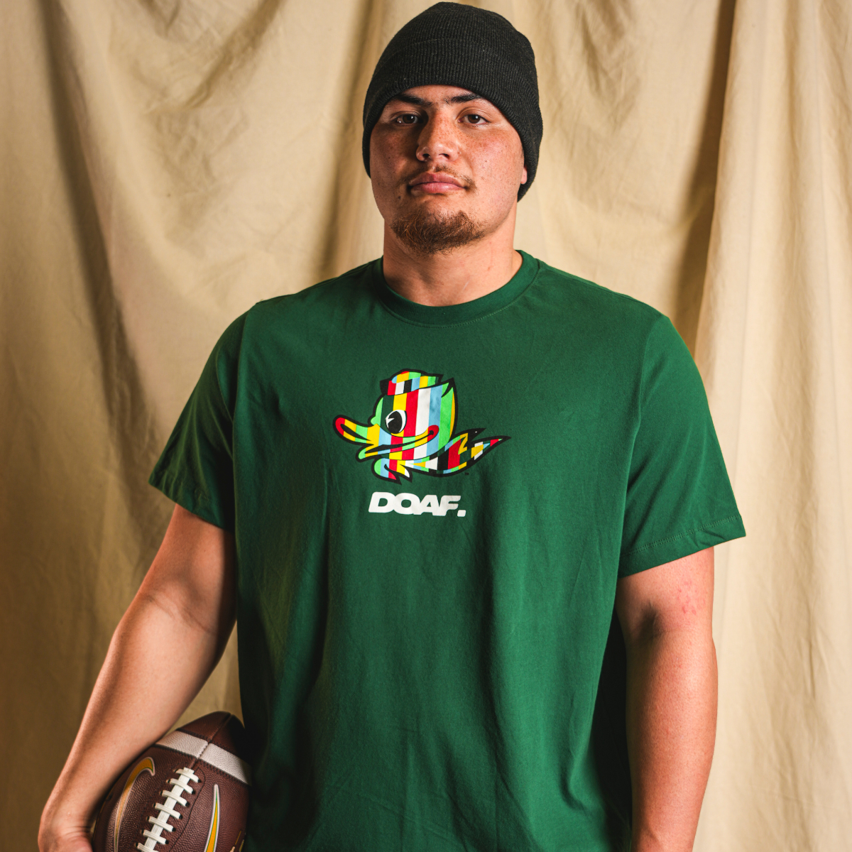 Teitum Tuioti wearing a green DOAF t-shirt with a colorful Duck graphic and text, holding a football against a beige curtain background.