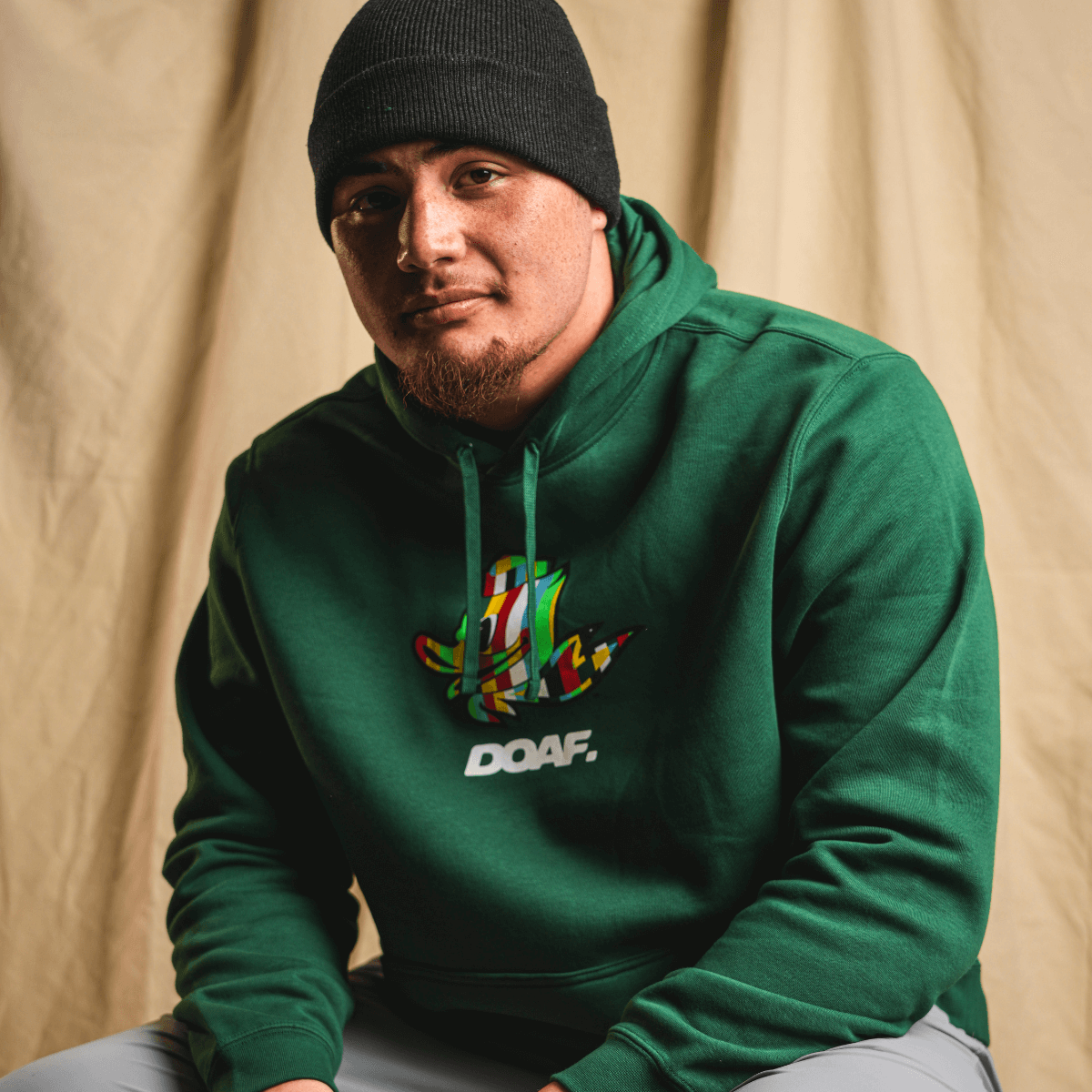 Teitum Tuioti wearing a green hoodie with a colorful logo and 'DOAF' text against a beige background