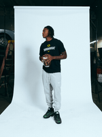 Dakorien Moore wearing a black DOAF Speedway t-shirt  with wellow logos and gray Speedway pants, standing against a white backdrop while holding a football.