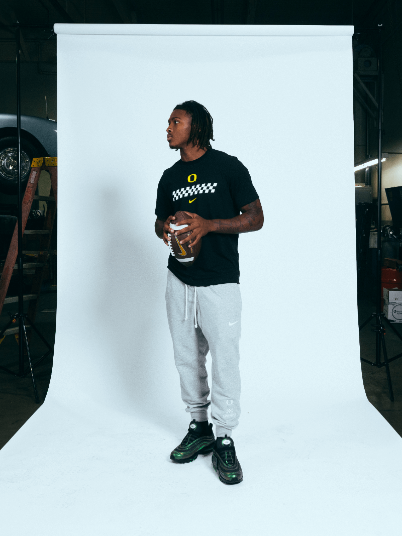 Dakorien Moore wearing a black DOAF Speedway t-shirt  with wellow logos and gray Speedway pants, standing against a white backdrop while holding a football.