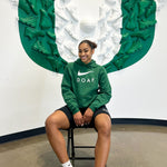 Deja Kelley wearing a green hoodie with 'DOAF' text, sitting in front of a large green and white  U of O logo.