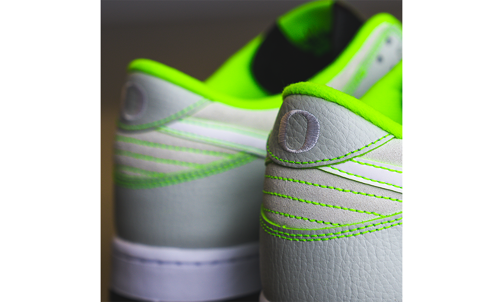 Close-up of gray sneakers with green accents on a white background. The DOAF Nike Dunk Low “Oregon” Player Edition, offers a fresh take on a classic shoe, with subtle interplays of texture and color highlighted by distinctive signature touches. The shoe features overlays of light grey tumbled leather accented by electric green stitching.