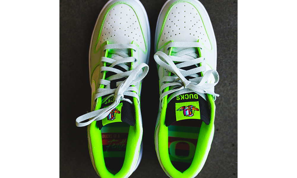 The Nike Dunk Low “Oregon” Player Edition. The shoe features a subtle tone-on-tone combination of off-white split suede quarter panels and toe boxes, and overlays of light grey tumbled leather accented by electric green stitching throughout, which also outline the white leather Swoosh branding. Additional branding, as well as another color pop, are provided by an electric green tongue tag sporting a multi-colored Duck head logo over all caps “DUCKS” branding.
