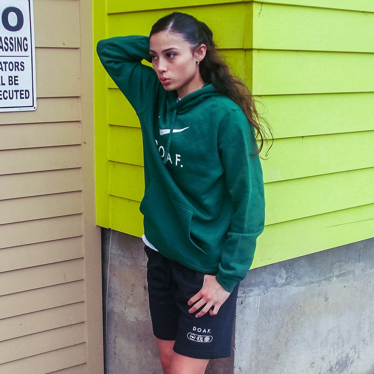 Katie Fiso wearing a set of DOAF gear, including black world wide webbed shorts,  against a green wall.
