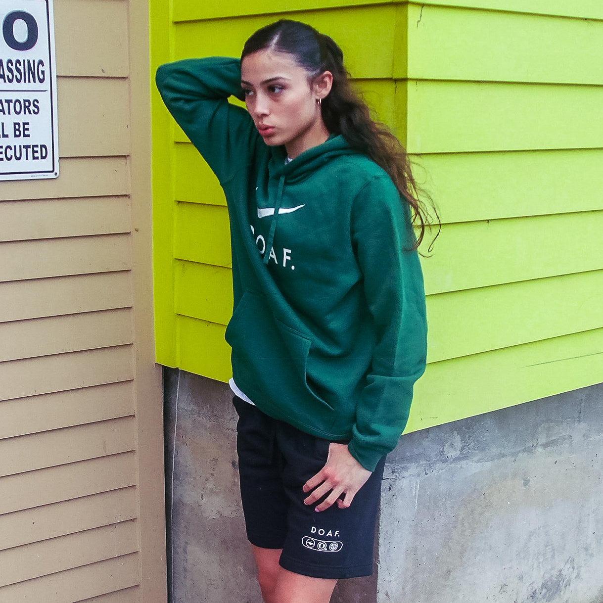 Katie Fiso wearing a set of DOAF gear, including black world wide webbed shorts,  against a green wall.